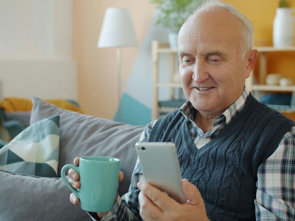 Lachende oudere man op de bank met een mok thee en zijn telefoon (illustratiefoto)