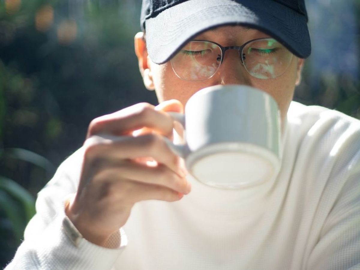 Man met pet en bril die uit een witte kop drinkt (illustratiefoto)